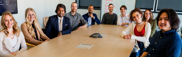 Diverse people of various genders and backgrounds sit around a board table smiling.