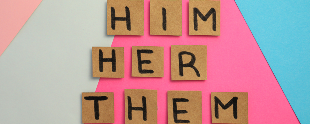 Bits of cardboard laid out to form words on a background in the colours of the trans flag. The words being formed are the pronouns ‘he’, ‘her’, ‘them’.