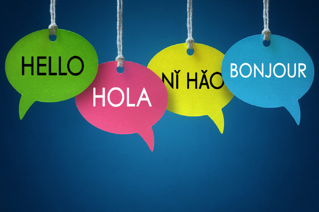 Four hanging speech bubbles on a blue background display "Hello," "Hola," "Nǐ Hǎo," and "Bonjour" in green, pink, yellow, and blue.